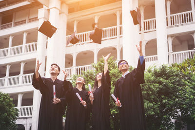 Students in university gowns throwing mortarboards while standing against building