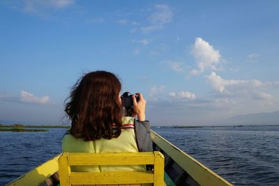 Rear view of woman photographing sea while sitting in boat against sky