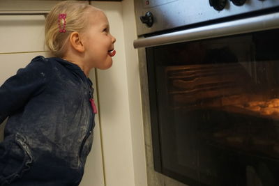 Side view of girl sticking out tongue while looking into oven at home