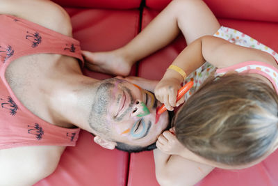 Top view of adorable girl painting face of calm father while relaxing together on sofa and having fun during weekend