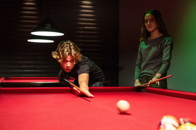Teenage girl standing by boy playing pool on illuminated red table