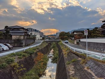 Road and canal by buildings against sky during sunset