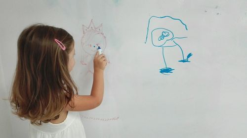 Rear view of girl holding paper while standing against white wall