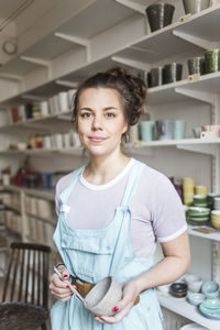 Portrait of confident young female potter holding hand tool and vase while standing against shelves at store