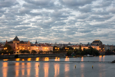 Illuminated buildings by river against cloudy sky