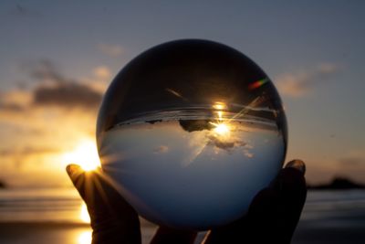 Cropped hand of woman holding crystal ball at beach against sky during sunset