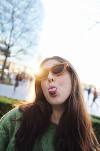 Young woman wearing sunglasses sticking out tongue in front of bright clear sky