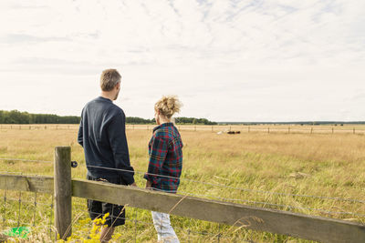 Rear view of couple standing by fence on grassy field against sky