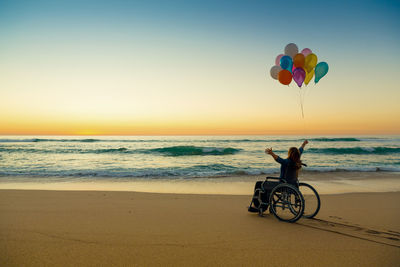 Silhouette mid adult woman with arms raised holding colorful balloons while sitting on wheelchair at beach during sunset