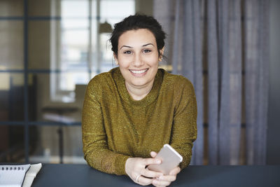 Portrait of smiling businesswoman sitting with mobile phone at desk in creative office