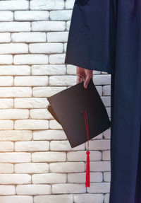 Midsection of person holding mortarboard against brick wall