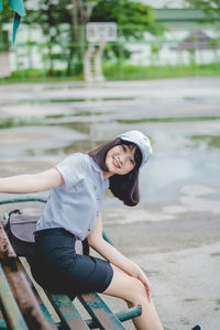 Portrait of smiling young woman sitting on bench in park during rainy season