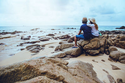 Rear view of men sitting on rock at beach against sky