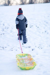 Rear view of boy with sled walking on snowy land