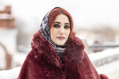 A slavic woman in a national colored scarf,a fur coat and  mittens on the porch of a wooden house