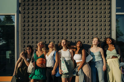 Smiling teenage girls with eyes closed enjoying sunny day while standing against wall