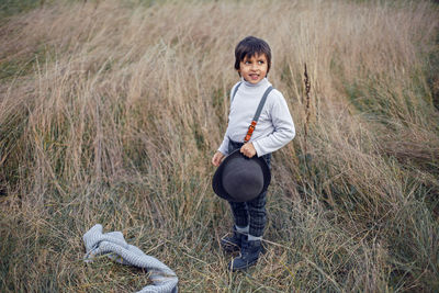 Boy child in plaid pants, hat, suspenders and scarf stands in a field in autumn