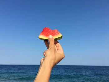 Cropped hand holding watermelon slice by sea against clear blue sky