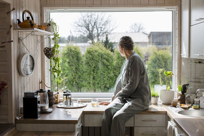 Mature woman sitting on kitchen counter at home looking out of window