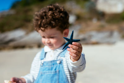 Soft focus of blue sea star in hand of adorable preschool boy with curly hair strolling on sunlit beach in summer