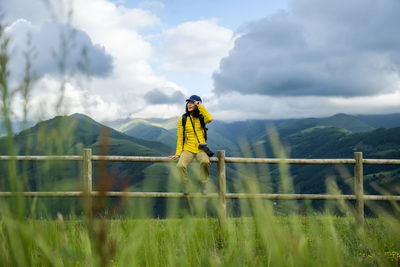 Portrait of beautiful asian girl sitting on handrail with green hills and valley behind her and having rest after hiking with her camera