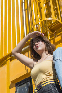 Low angle view of young woman shielding eyes while standing against building