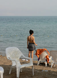 Rear view of woman standing by chairs at sea shore against sky