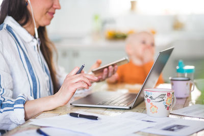 Midsection of working mother using technologies while daughter sitting in background at dining table