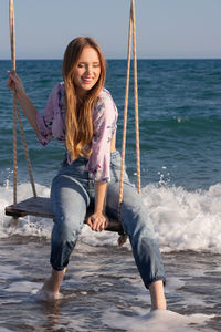 Portrait of smiling young woman against sea sitting on the swing