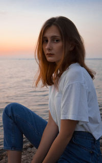 Beautiful young woman sitting on sea shore against sky during sunset