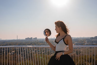 Woman holding mirror in city against clear sky