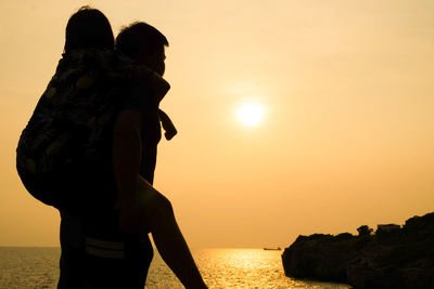 Man giving piggyback ride to sister by sea against orange sky