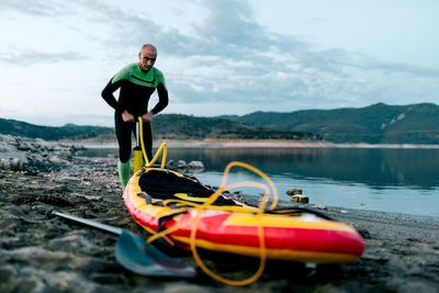 Male surfer in wetsuit pumping sup board while standing on seashore and preparing for surfing