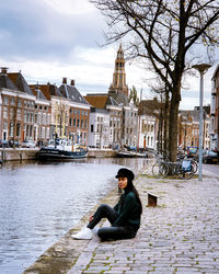 Full length of woman sitting by canal against buildings and sky