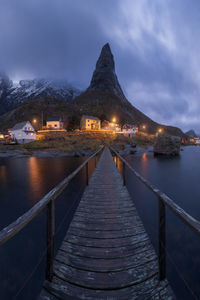 Amazing view of wooden footbridge crossing calm river and leading to illuminated village with cottages located at bottom of highlands in reine , lofoten islands in norway in evening