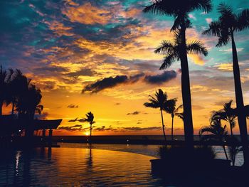 Silhouette palm trees by swimming pool against sky during sunset
