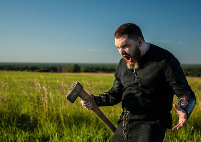 Angry man with axe standing on grassy field against sky
