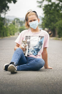 One young girl or woman wearing face mask seated on street carrying a young white and gray cat in a blurred natural environment