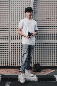 Full length of young man holding camera while standing on footpath