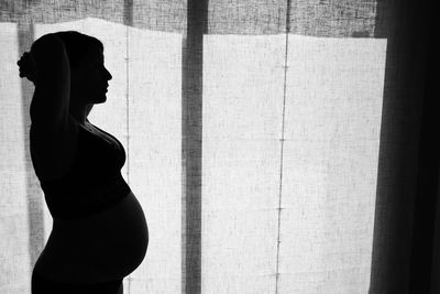 Side view of silhouette pregnant woman standing by curtain