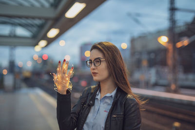 Young woman holding illuminated string lights while standing at railroad station platform at dusk