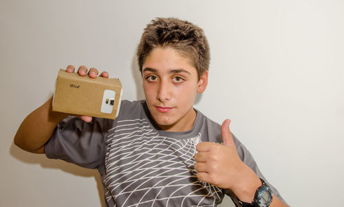 Portrait of boy holding virtual reality while gesturing thumbs up sign against gray background