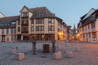 Illuminated street amidst buildings against sky at dusk