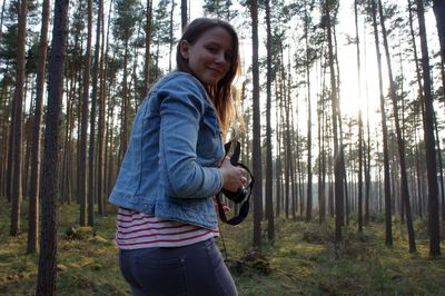 Portrait of smiling woman holding camera while standing against trees in forest