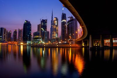 Illuminated modern buildings by river against sky in city at night