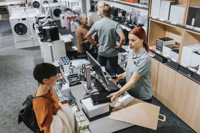 Mature woman standing at checkout counter while female sales clerk scanning appliances in store