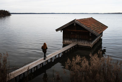 Unrecognizable female traveler in warm clothes standing on wooden plank pier near shabby shed located at lake in gloomy autumn day