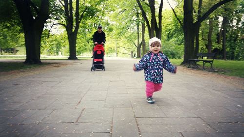 Portrait of cute girl running on footpath against father with baby carriage in park