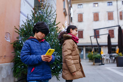 Two standing children on the street watching mobile phone next to decorated christmas tree