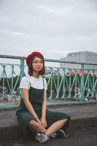 Portrait of young woman sitting on footpath in city against cloudy sky